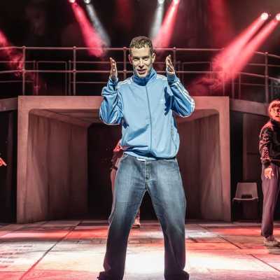 A young man in a pale blue tracksuit top stands centre-stage, gesturing with his hands forwards. A tunnel is behind him, with a curved bridge over it, and there are red stage lights. 