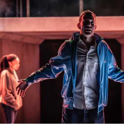 A man, wearing a blue tracksuit top, white shirt and blue jeans stands on a stage. His mouth is slightly open, face slightly hidden behind shadows from the stage lights, and his arms are out to the sides. Behind him on the left, out of focus, is a woman wearing a hoodie and jeans.  