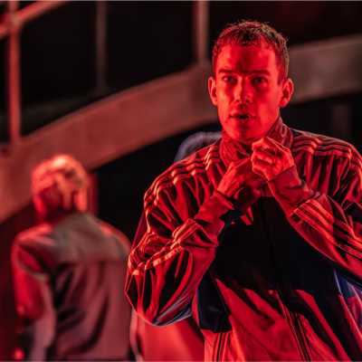 A half-body shot of a man wearing an Adidas tracksuit top. He is staring intensely, with his mouth slightly open. His hands are on the zip of his top, as if doing it up. The image has a red hue from the stage lighting.  