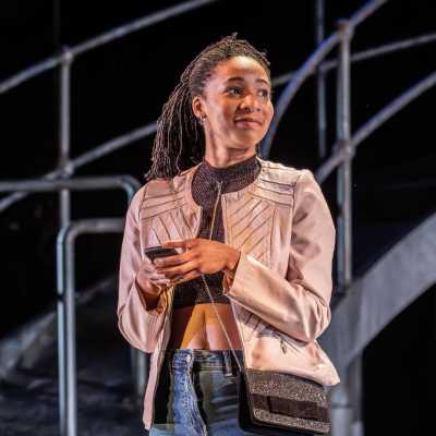 A woman, wearing a beige jacket, brown top, blue jeans and a sparkly handbag stands on a stage. She is holding a mobile phone in both hands and is looking to her left with a small smile on her face. Behind her is a metal bridge. 