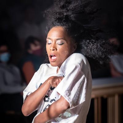 Anoushka Lucas as Laurey Williams, singing and with arms wrapped around self. Members of the audience are visible in the background.