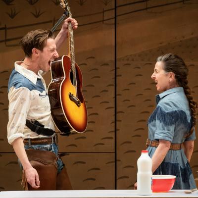 Arthur Darvill as Curly McLain holding a guitar. Standing opposite is Liza Sadovy as Aunt Eller, both are singing.