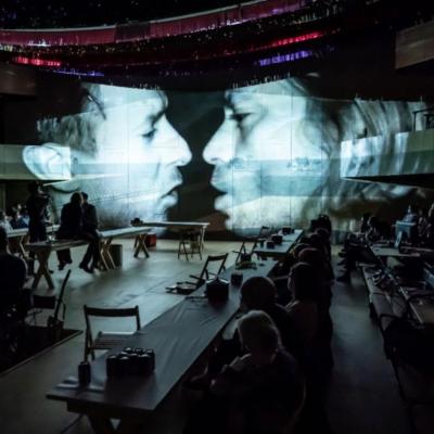 An auditorium of people in low light, with the silouhettes of multiple figures visible. A projection on the wall shows the faces of Patrick Vaill as Jud Fry and Arthur Darvill as Curly McLain about to meet in near darkness.