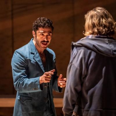 Stavros Demetraki, as Ali Hakim, is wearing a denim jacket and gold chains and rings; Patrick Vaill as Jud Fry is standing wearing a dark blue hoodie and t-shirt, with back to the camera