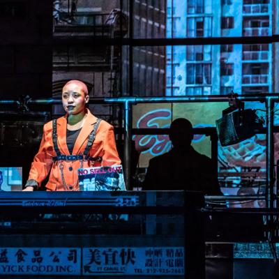 XANA in The Collaboration (c) Marc Brenner. A DJ, in an orange suit and leather harness, stood in a DJ booth with a projection on New York behind them